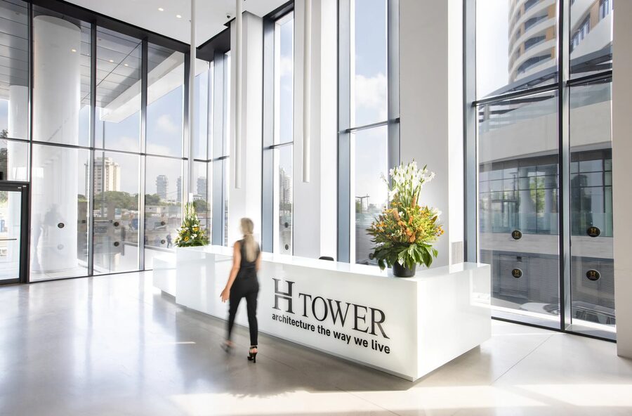 דלפק קבלה לבן עם השלט H TOWER, סידורי פרחים וחזית זכוכית; אישה חוצה את הלובי