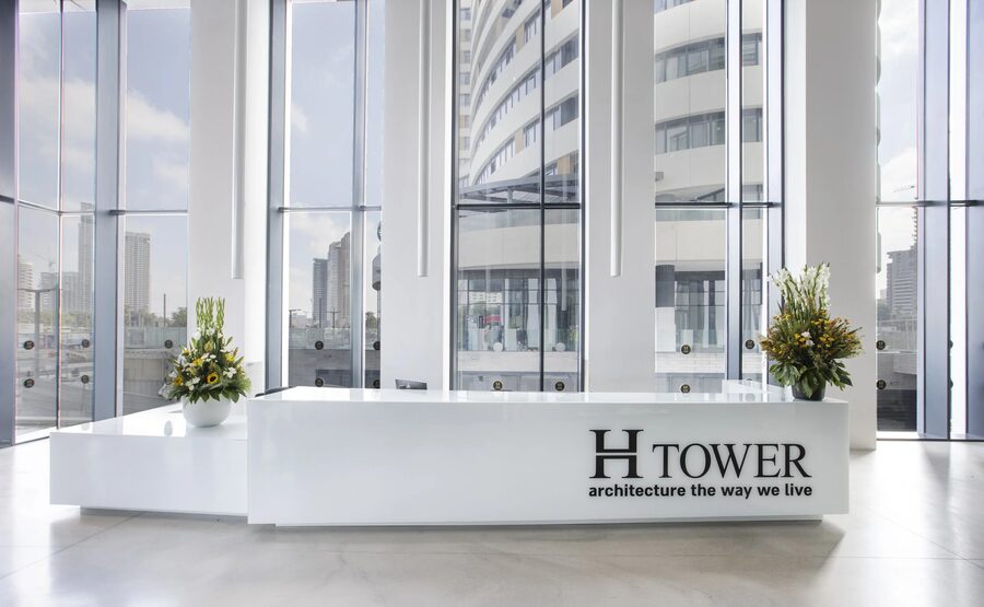 דלפק הקבלה עם השלט H TOWER, שני סידורי פרחים וחזית זכוכית הפונה אל מגדלי העיר